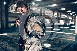 A picture of military man posing on camera. He is in a big hangar. He is holding black rifle and ready to shoot. Guy is