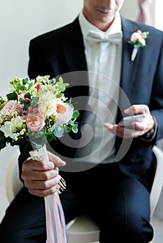 Picture of a modern broom in his finest look texting on his phone holding a cute wedding bouquete photo