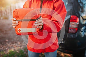 Picture of orange firt aid kit bag young oman hold with both hands. She stads at tent. Model wears orange jacket.