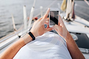 Picture of phone in man`s hands. He holds in confident. Screen is dark. Phone is white. Young man lies on yacht board.