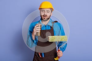 Picture portrait of sleepy tired construction worker closing his eyes, holding thermo mug and roller in both hands, wearing brown