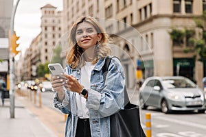 Picture of pretty young woman staying on the street holding phone in hands.