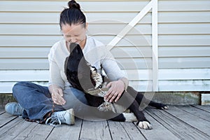 Picture of seated woman in jeans and white jacket stroking black dog on white background wooden wall