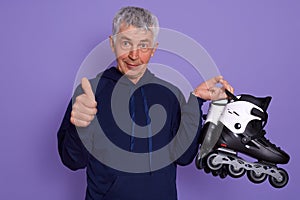 Picture of senior man wearing sporty clothing, holding roller skates, showing thumb up, being happy after active rollerblading in