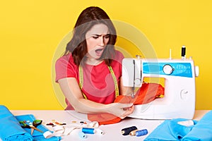 Picture of shocked desperate young inexperienced seamstress working with red fabric, having troubles with sewing, having