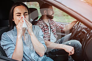 A picture of sick girl riding in car with young man. She is sneezing in napkin while he is paying attention to the road photo