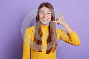 Picture of smiling lady looking directly at camera, standing against lilac studio wall and showing phone gesture with fingers.