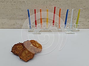 A glass Hannuka filled with colourful candles on a white table with three fritters and a stone wall in the background outdoor