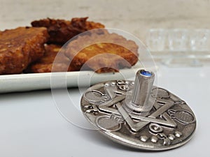 A plate full of Hannuka potato latkes fritters with a silver dreidel on a white table with a glass chanukiah in the background