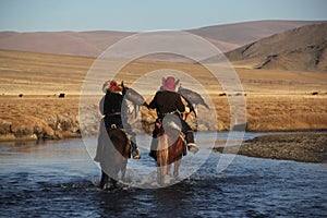 Picture of two horseriders in a river surrounded by a valley with hills on a blurry background