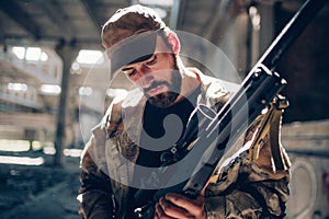 A picture of warrior standing in a hangar and looking down at his rifle. He is cleaning it and prepare it to fight