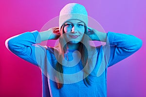 Picture of young adorable girl wearing sweater and cap, standing isolated over pink neon background, looking at camera with