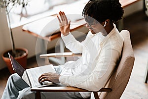 Picture of young afro man talking by videocall by laptop computer and waving indoors