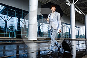 Picture of young redhaired businessman holding black umbrella and suitcase walking in rain at airport