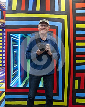 75 year-old Caucasian tourist posing before a mirror and taking a picture of his reflection at Wonderland Dreams in New York City.