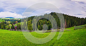 Picturesque spring Carpathians scene with wooden split rail fence across a green and lush pasture, and old cabins on the valley