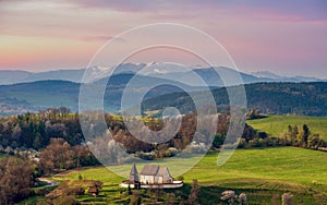 Picturesque spring mountain landscape with hills, meadows and romantic historic wooden building at colorful sunset.