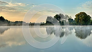 A picturesque sunrise over a foggy lake with trees on the shore reflected in the water. Morning of the end of summer, beginning of