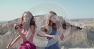 Picturesque view of female violinists duet in blowing dresses play among cliffs