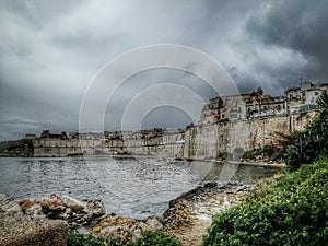 Picturesque view of the wall of old town and houses on the hill on the coast under cloudy sky, Malta