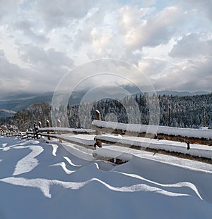 Picturesque waved shadows on snow from wood fence. Alpine mountain winter hamlet outskirts, snowy path, fir forest on far misty