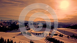 Picturesque white mountain slopes covered with pine forests and skiing pistes and moving chairlift in eavning Sunset aerial view.