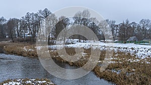 Picturesque winter or late autumn nature landscape with river, trees, dried yellow grass at cloudy frosty day.