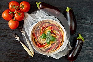 Pie with eggplants and tomatoes in the baking dish decorated with fresh basil leaves. Homemade vegetable tart on the black table