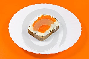 A piece of black rye bread on a white plate smeared with butter and red caviar