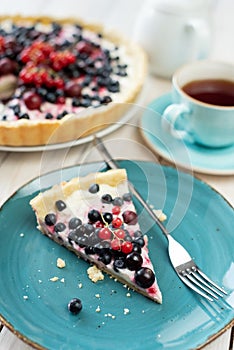 Piece of blueberry open pie on blue plate with cup of black tea and white tea pot on bright painted wood background