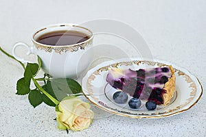 Piece of blueberry pie on a plate, cup of tea and white rose on a white background - close up