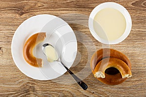 Piece of bread rings, spoon with condensed milk in plate, bowl with milk, baranka on wooden table. Top view photo