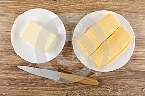 Piece of butter in saucer, plate with slices of cheese, knife on wooden table. Top view