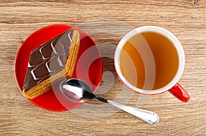 Piece of cake in red saucer, spoon, cup of tea on wooden table. Top view
