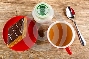 Piece of cake in red saucer, sugar bowl, spoon, cup of tea on wooden table. Top view