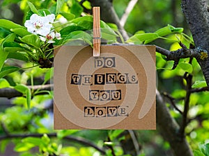 Piece of cardboard with words Do Things You Love on it hanging on a pear tree branch with blossoms and leaves using a wooden