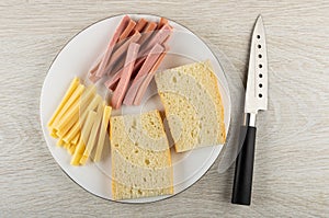 Piece of cheese, sausages, slices of bread in white plate, knife on table. Top view