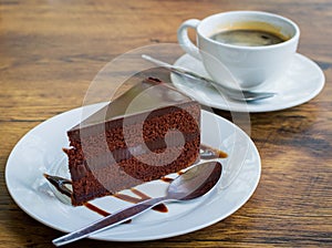 Piece of chocolate cake with spoon on white plate. Cup of coffee on background