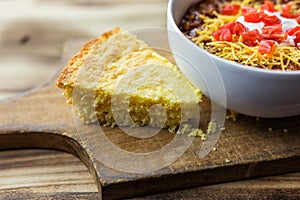 A piece of cornbread on a vintage cutting board shown with a bowl of chili.
