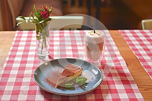 Piece of cream chocolate cake with cacao powder and mint served with Latte coffee on a table with red plaid tablecloth.