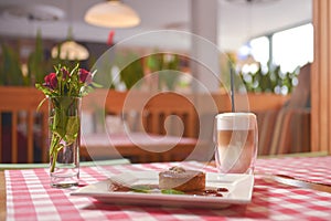 Piece of cream chocolate cake with cacao powder and mint served with Latte coffee on a table with red plaid tablecloth.