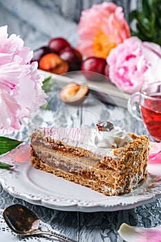 Piece of delicious cake with cream and plum jam on the plate over grey background with fresh plums and peonies flowers. Sweets,