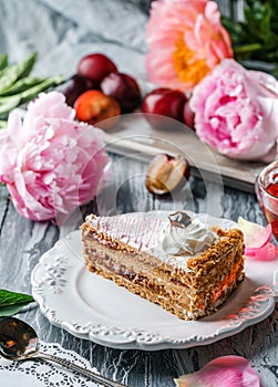Piece of delicious cake with cream and plum jam on the plate over grey background with fresh plums and peonies flowers. Sweets,