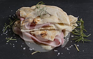 A piece of focaccia is garnished with slices of cheese and ham, decorated with sprigs of rosemary and coarse salt, on a slate base