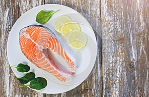 A piece of fresh salmon fish steak on a white plate with spinach and lemon, on  a gray wooden background. Omega 3 vitamin, healthy