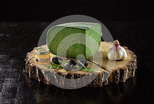 Piece of green pesto cheese, jar of jam, olives and garlic on wooden board