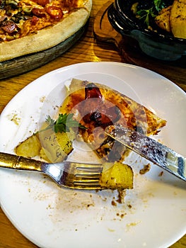 A piece of half-eaten pizza on a white plate with  fork and knife. Wooden plate with a pizza and a bowl of baked potatoes