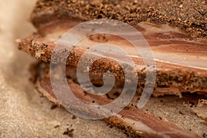 A piece of homemade bacon with pepper on white oiled paper, close-up, selective focus