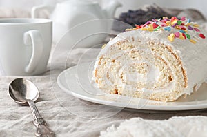 A piece of homemade biscuit roll with butter cream. Decorated with colorful topping. On a white plate. In the background is a