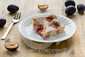 Piece of homemade plum pie on a white plate with organic plums and fork on wooden table